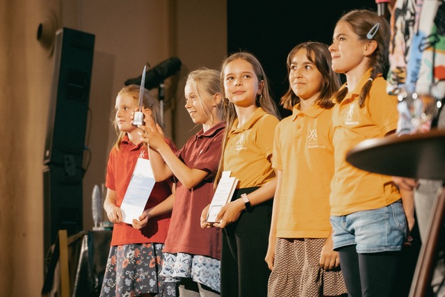 Fünf Kinder in bunten T-Shirts halten eine Trophäe in Form einer Feder und Urkunden hoch.